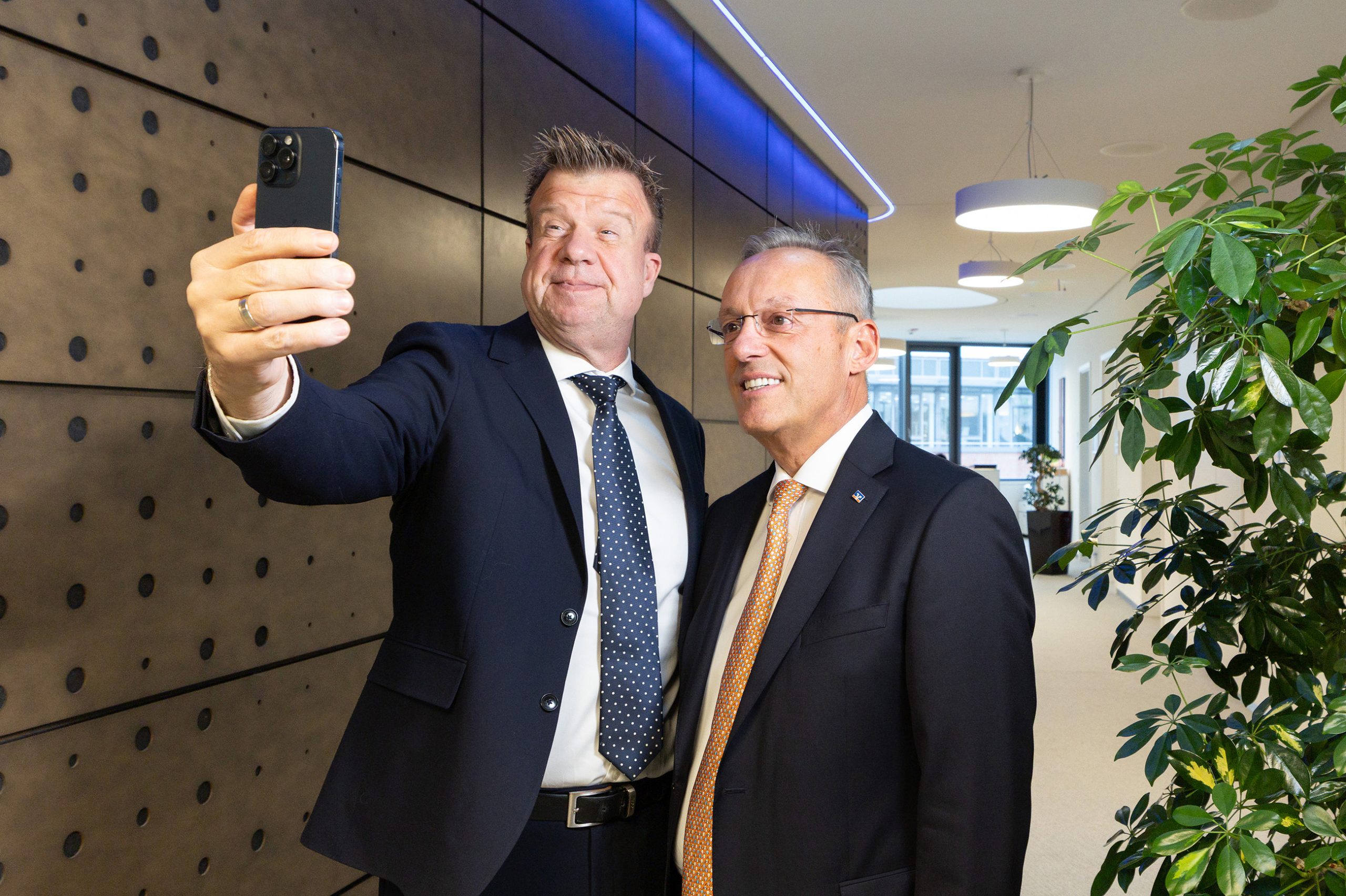 Unsere Vorstandssprecher in einem modernen Umfeld der Volksbank Mittelhessen, nah beieinander stehend. Sie halten ein Smartphone hoch, mit dem sie gemeinsam ein Selfie aufnehmen.