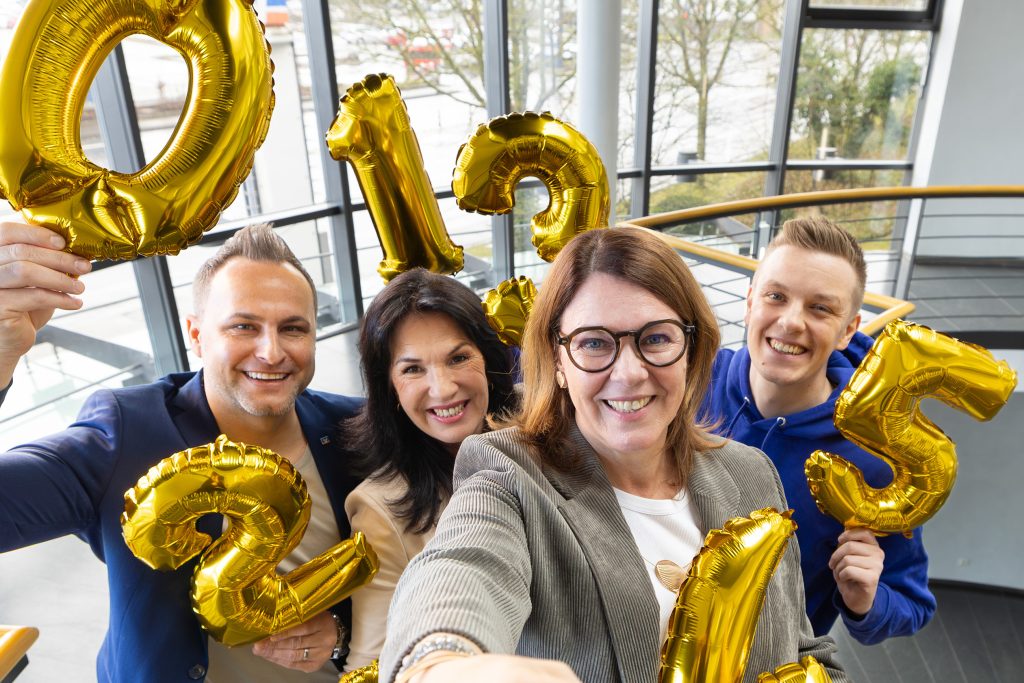 Gruppenfoto auf einer hellen Innentreppe mit großer Fensterfront. Vier Personen stehen eng zusammen und halten goldene Folienballons in Form der Zahlen 0, 1, 2, 3, 4 und 5 in die Kamera. Die Ballons dominieren das Bild.