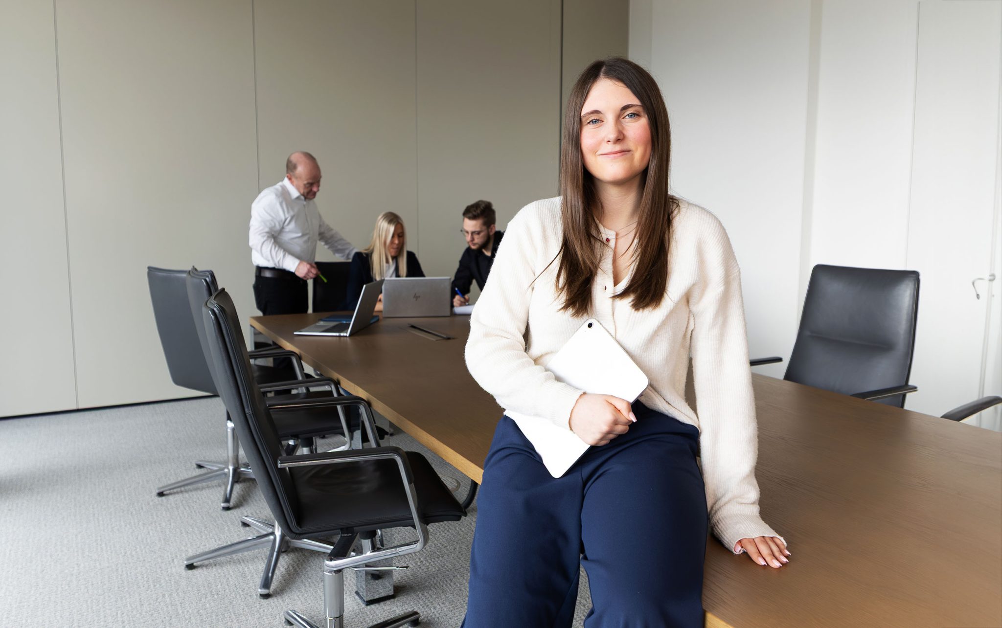 Eine Auszubildende sitzt im Vordergrund auf der Tischkante, sie hält ein Tablet im Arm. Im Hintergrund unterhalten sich drei ihrer Kollegen.