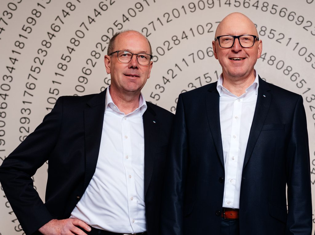 Zwei Generalbevollmächtigten der Volksbank Mittelhessen stehen in dunklen Business‑Anzügen vor einer Wand mit kreisförmig angeordneten Zahlen.