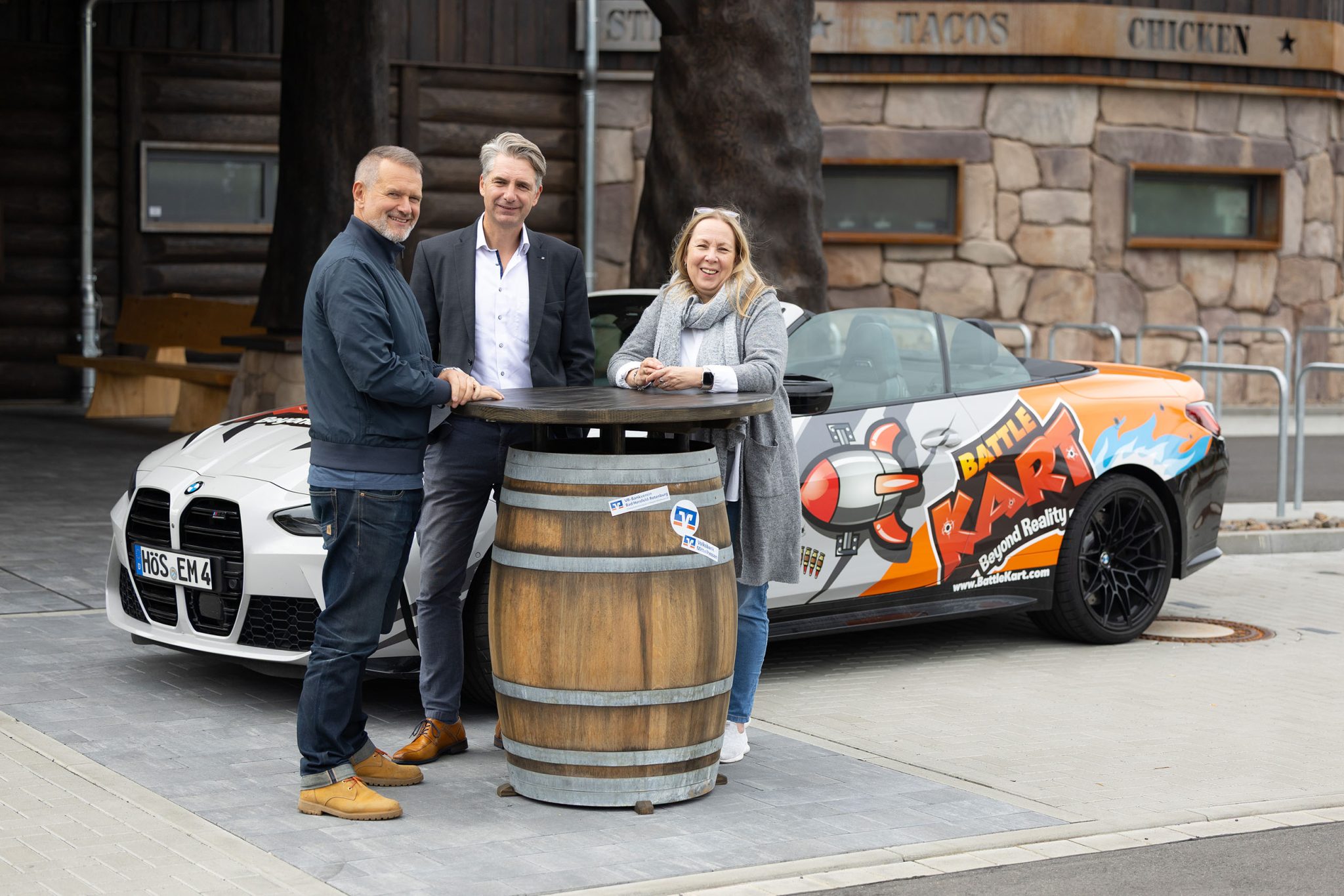 Die Unternehmer von Timberjacks und BattleKart stehen mit einem Firmenkundenberater der Volksbank Mittelhessen an einem hohen Fass‑Tisch vor einem bunt gestalteten BattleKart‑Fahrzeug und dem im Blockhausstil errichteten Timberjacks‑Restaurant.