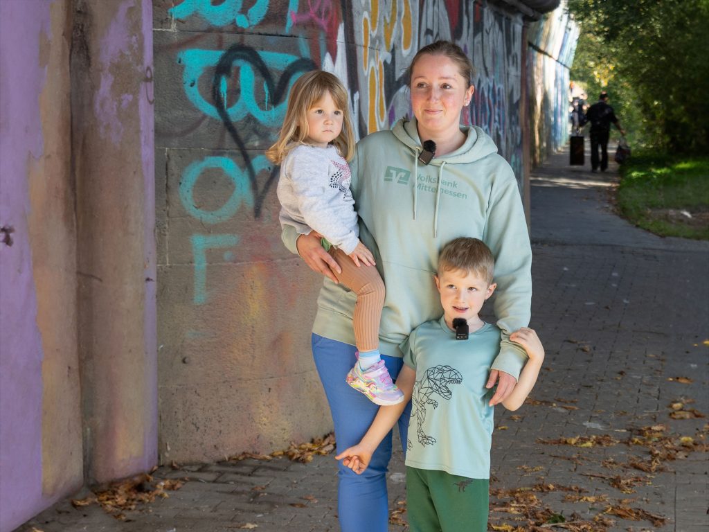 Eine Mitarbeiterin der Volksbank Mittelhessen steht mit ihren zwei Kindern bei der Lahnreinigung unter einer bunt besprühten Brücke.
