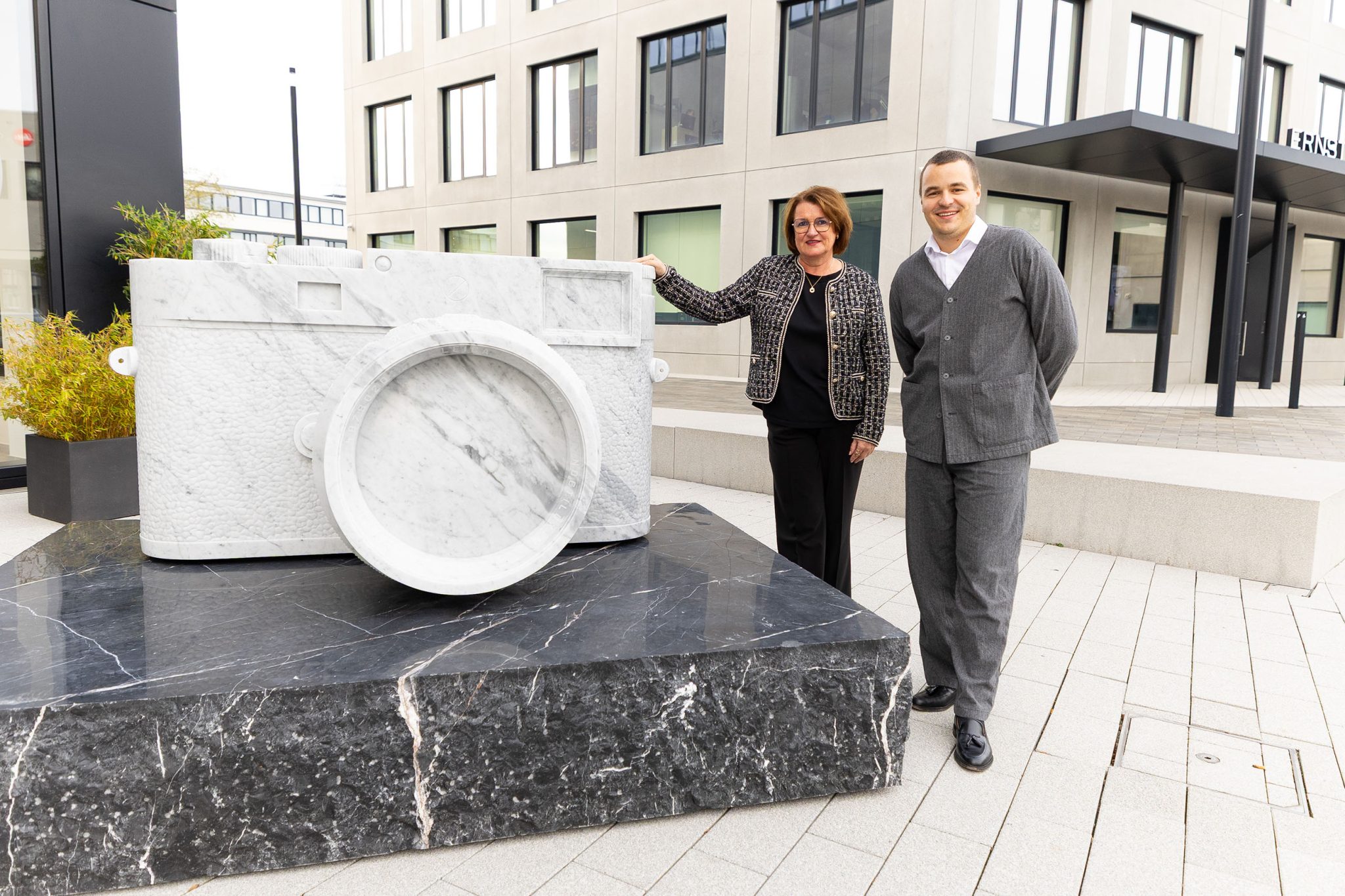 Zwei Führungspersonen des Unternehmens stehen vor einer großen weißen Marmorskulptur in Form einer Leica‑Kamera auf dem Außengelände des Leitz-Parks in Wetzlar.
