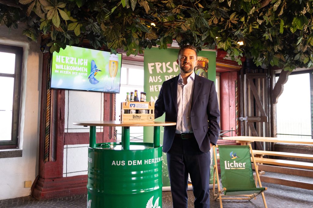 Ein Firmenkundenberater der Volksbank Mittelhessen besucht die Licher Privatbrauerei und steht an einem Promotion‑Stehtisch mit Licher‑Bierpräsentation.