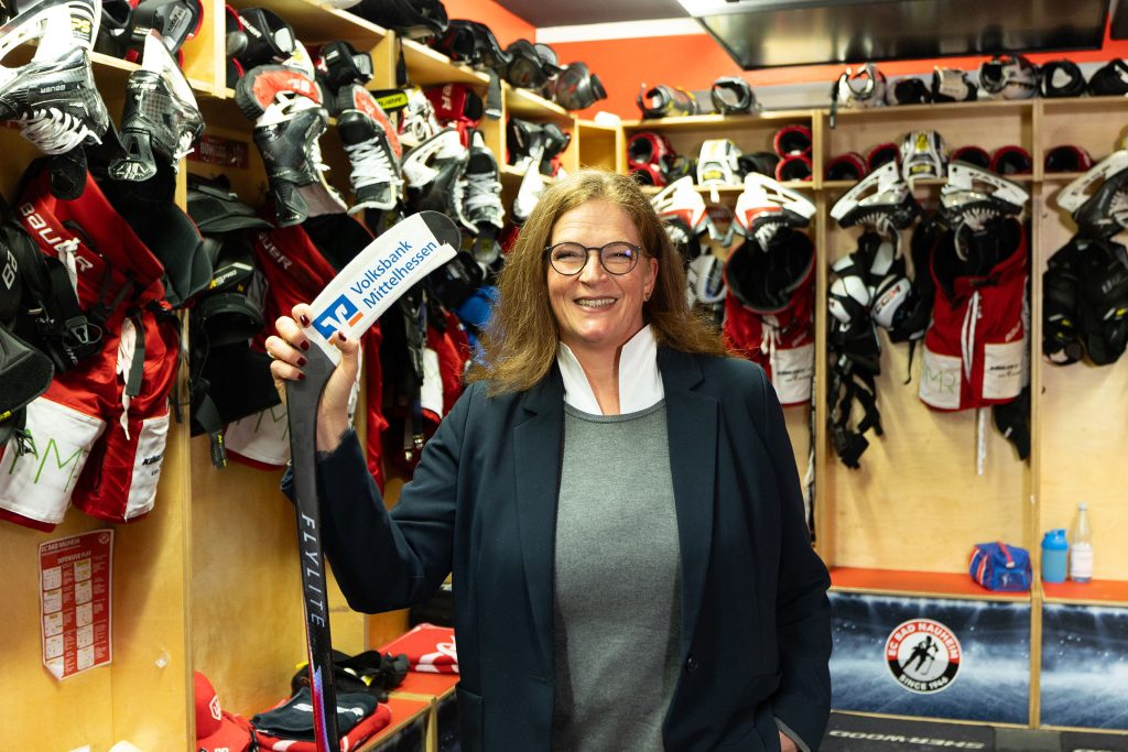 Eine Firmenkundenberaterin der Volksbank Mittelhessen hält einen Eishockeyschläger in der Kabine des EC Bad Nauheim und besucht den Verein vor Ort.