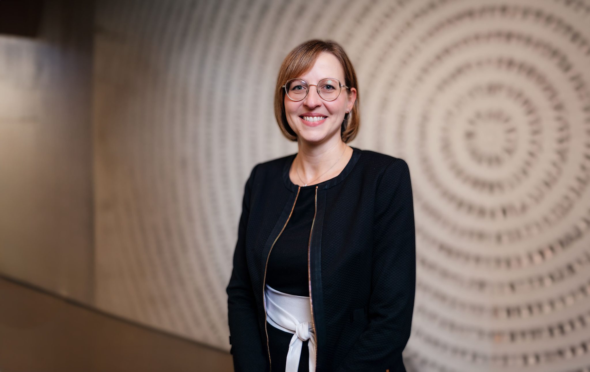 Eine Generalbevollmächtigte der Volksbank Mittelhessen steht elegant gekleidet in einem modernen Innenbereich vor einer Wand mit kreisförmig angeordneten Zahlen.