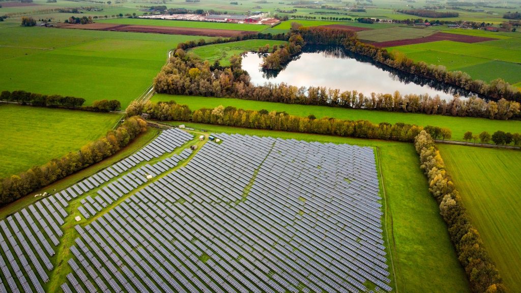 Luftaufnahme eines großen Solarparks mit dicht angeordneten Solarmodulen inmitten grüner Felder. Neben der Anlage liegen Baumstreifen und ein Gewässer; die Umgebung wirkt landwirtschaftlich geprägt und naturverträglich erschlossen.
